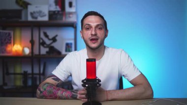 Dövmeli Avrupalı bir adam, gösterişli bir stüdyo ortamında podcast yaparken düşüncelerini paylaşıyor. Doğal ton ve temiz çerçeve.