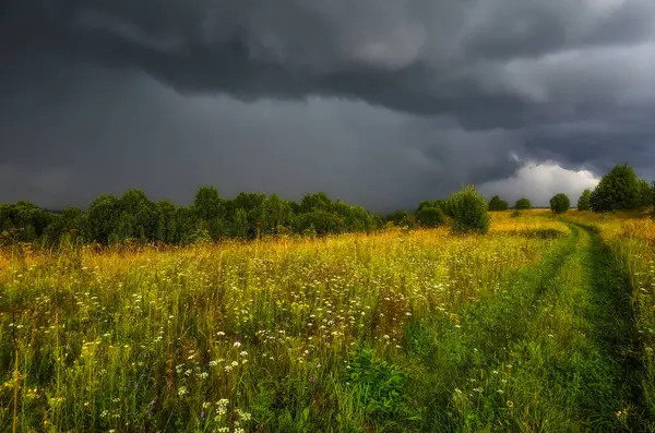 Dramatic sky with gray rainy clouds over summer meadow on hill before storm. Summer rural landscape with dirt road in rainy weather. Weather forecast concept