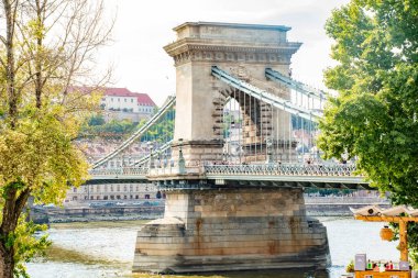 Bu fotoğraf Macaristan 'ın Budapeşte kentindeki Szechenyi Zinciri Köprüsü' nün uzak bir görüntüsünü sunuyor, bu ikonik simgenin ihtişamını ve genişliğini şehir manzaralı gökyüzüne yansıtıyor. Görüntü