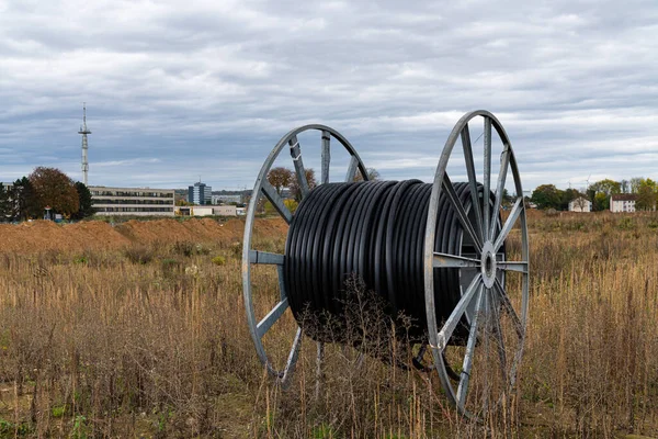 Large metal cable reel with black plastic cable on a vacant lot. The gray sky is covered with clouds.