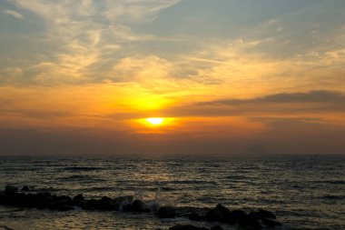 Denizin üzerindeki kara bulutlarda dramatik bir gün batımı. Dalgalar kayalara çarpar.