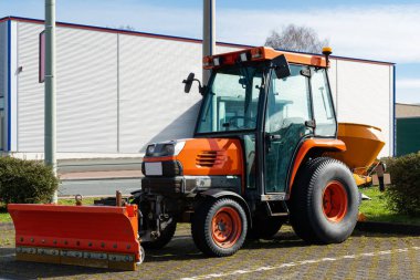 Küçük turuncu tekerlekli bir traktör bir otoparkta duruyor. Profil kağıdıyla kaplı bir binanın arka planına karşı..