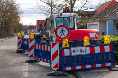 Bariyerlerin arkasında kaldırımda duran küçük bir traktör. Yaya trafik levhası yok. Kablo.