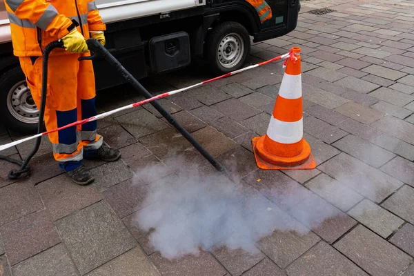 Road worker cleans the pavement with a steam pressure hose. Orange-white conical pylon of fencing tape.