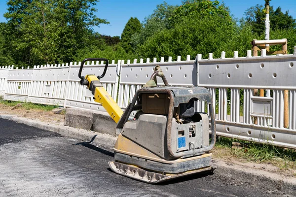Manual road tamping machine at a construction site in front of a plastic white fence.