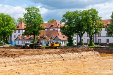 İnşaat alanındaki sarı kazıcı bir traktöre toprak yüklüyor. Arka planda kiremitli çatıları olan beyaz bina.