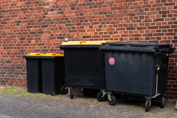 Black plastic garbage cans with yellow lids standing near the wall of a brick building.