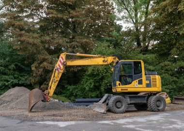 Yeşil ağaçların arka planına karşı bir çakıl yığınının önündeki sarı kazıcı. Kenar veiw.