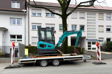 Üzerinde küçük bir kazıcı olan bir araba römorku yol kenarında bir apartmanın önünde duruyor..