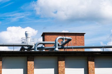 Beyaz bulutlu mavi gökyüzünün altında çeşitli boyutlarda havalandırma boruları olan endüstriyel kırmızı tuğla bir binanın çatısı..