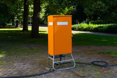 Parlak turuncu bir alet kutusu yeşil çimlerle çevrili bir parktaki metal bir standın üzerinde duruyor..