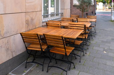 Bir sıra ahşap masa ve sandalye bir restoran veya kafenin dışındaki kaldırım taşı bir sokakta duruyor. Masalar boş sandalyelerle dolu, misafirleri karşılamaya hazır. Sıcak güneş ışığı ve büyüleyici