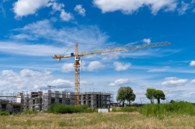 Sarı bir vinç, beyaz bulutlu mavi bir gökyüzüne karşı duruyor. İnşaat halindeki yeni bir binanın üzerine uzanıyor..