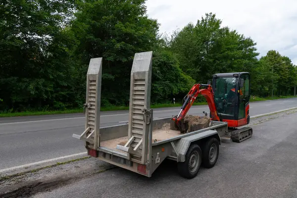 Kırmızı bir kazıcı yol kenarındaki bir karavana yükleniyor, nakledilmeye hazır. Karavanın aşağıda iki rampası var ve kazıcı kovası yerde dinleniyor..