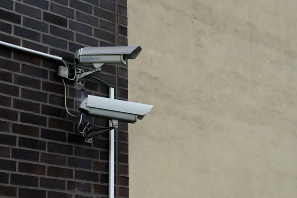 İki güvenlik kamerası bir tuğla duvara monte edilmiş, biri diğerinin üstünde. Kameralar dışarıyı gösteriyor, çevreyi yakalıyorlar..