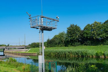 Platformu ve güvenlik kameraları olan metal bir kule, arka planda köprüsü olan kırsal bir alanda bir kanala bakıyor..
