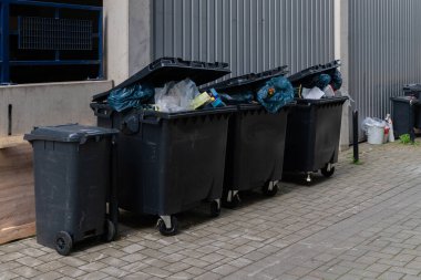 Kentsel bir ortamda, ticari bir binanın önünde içi atık dolu bir sürü çöp kutusu sıraya dizildi..