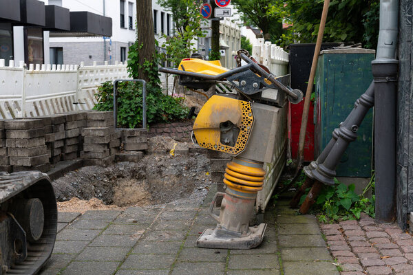 A close-up of a construction site showcasing a vibratory rammer positioned next to a pit, with stacked paving stones and safety barriers in the background..