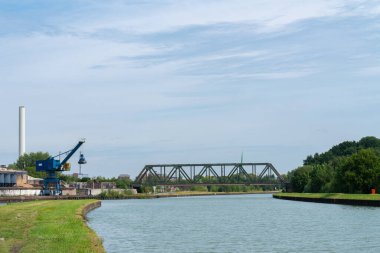 Berrak mavi gökyüzünün altında bir vinç ve köprünün yer aldığı sanayi bölgesinin yanından akan sakin bir nehir doğa ve ticaretin bir karışımını sergiliyor..