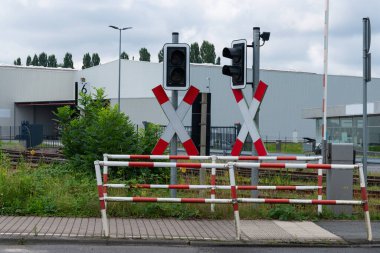 Bulutlu bir yaz gününde endüstriyel bir bölgede rayların olduğunu gösteren iki demiryolu geçidi sinyali beyaz bir bariyerin yanında duruyor..