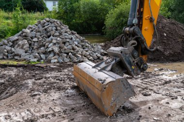 Bir inşaat sahasına büyük bir kazıcı yerleştiriliyor. Etrafı kayalar ve yeşilliklerle çevriliyken toprağı hareket ettiriyor..