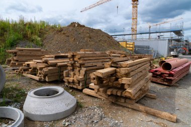 Sağlam ahşap kirişler ve borular bir inşaat sahasına düzgünce istiflenmiştir. Arka planda kirli bir tümsek ve bulutlu bir gökyüzünün altında vinçler vardır..
