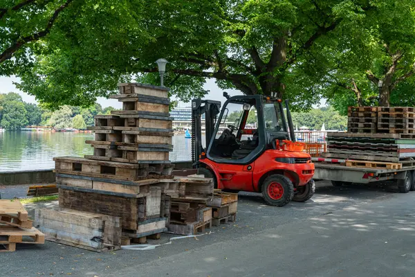 Bir forklift, sakin bir gölün yanında ahşap paletler taşır. Etrafı yeşillik ve açık hava aktiviteleriyle çevrilidir..