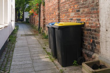 Dar bir sokakta renkli kapaklı plastik çöp bidonları tuğla bir duvara karşı dizilmişti..