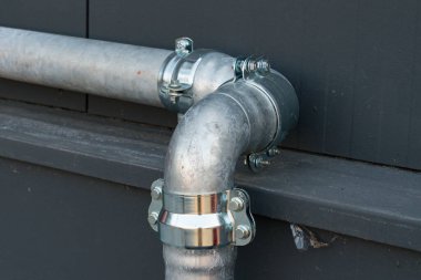 A close view of a metal plumbing pipe featuring a shiny elbow joint, attached securely to a building's exterior wall under bright daylight, showcasing the craftsmanship of plumbing installation. clipart