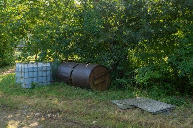 Paslı bir fıçı da dahil olmak üzere çok sayıda konteynır, ağaçlar ve çalılıklarla çevrili yemyeşil bir alandaki çakıl yolunun yanına bırakılarak ihmal hissi uyandırılıyor..