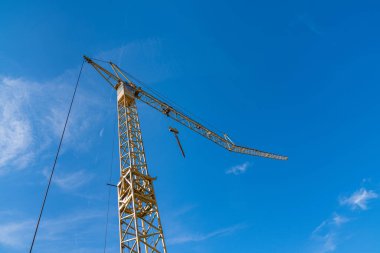 Uzun bir vinç açık mavi bir gökyüzüne karşı duruyor, yapısını gösteriyor ve aşağıdaki inşaat alanında aktif bir iş gününe hazırlanıyor..