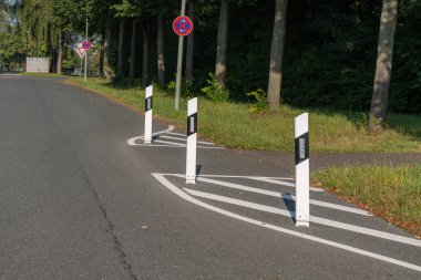 Trafik direkleri virajlı bir yolda, araçları viraja doğru yönlendiriyor. Bölge, kentsel ortam için doğal bir zemin oluşturan yemyeşil bir alana sahip..