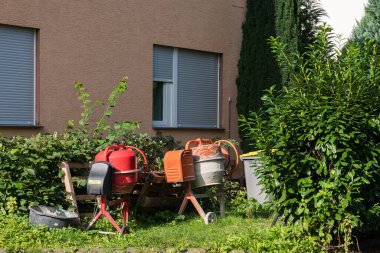İki çimento karıştırıcı, biri kırmızı ve biri gri, bir evin dışındaki çimenli bir bahçede oturuyorlar. Arka planda çalılar ve ağaçlar var..