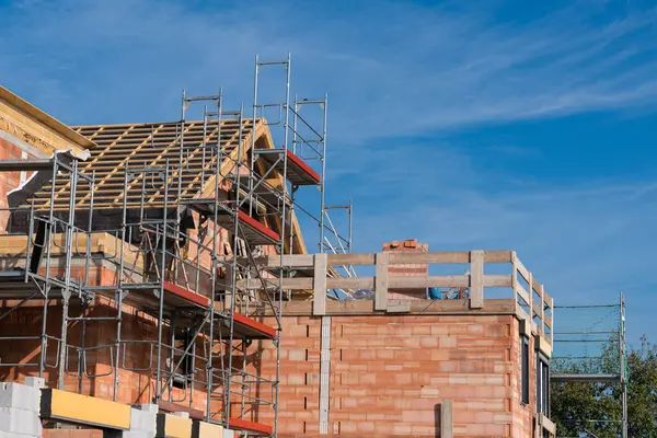 Bir inşaat sahasında tuğla duvarlara bağlı iskeleler sergileniyor. İşçiler, parlak mavi gökyüzü altında devam eden inşaat faaliyetlerini vurgulamak için inşaat süreciyle meşgul..