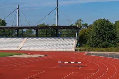 İyi bakımlı bir atletizm pisti, arka planda açık mavi gökyüzünün altında boş tribünlerle bir kum çukuru ve engeller oluşturur ve açık hava sporları için mükemmel bir ortam yaratır..
