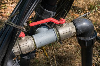 Sulama sistemi içindeki bir su vanası bağlantısının yakın plan görüntüsü, borular ve doğal çimen ve bitki örtüsü arasında kırmızı bir sapı var..