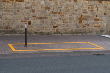 Turuncu boyayla çizilmiş boş bir park yeri sokak lambasıyla taş duvar arasında duruyor ve araçları parka davet ediyor. Bölge temiz ve bakımlı..