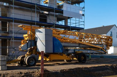 İnşaat vinci, inşaatın devam ettiği bir inşaat sahasına park edilmiş. İskeleyi çevreleyen yapı, açık mavi bir gökyüzü altında kısmen bitmiş yapıyı destekler.