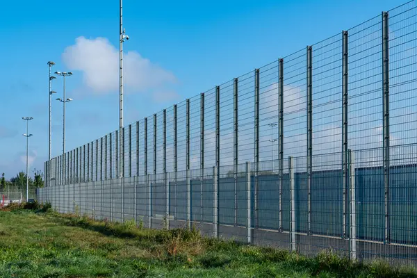Yüksek güvenlik çitleri parlak mavi bir gökyüzünün altında projektörlerle sabitlenmiş bir tesisi kapsıyor. Tabandaki yemyeşil otlar güvenli ortamı arttırıyor..