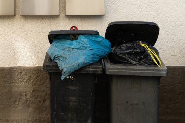 İki çöp bidonu yan yana duruyor, biri mavi bir çanta ve diğeri siyah bir çanta ile dolu, bölgedeki kentsel atık yönetimi sorununu yansıtıyor..