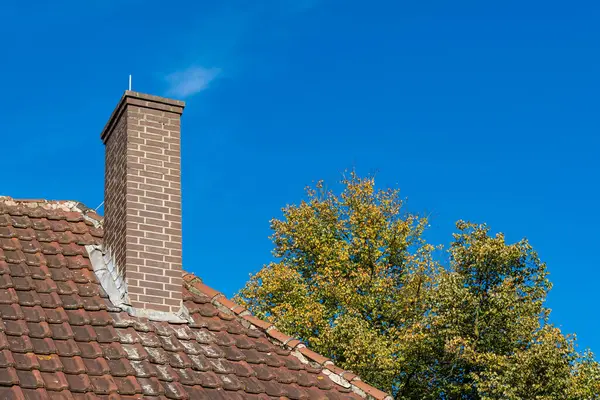 Bir tuğla baca, berrak mavi bir gökyüzüne karşı, yıpranmış bir çatıdan yükselir. Yakındaki bir ağaçtaki altın sonbahar yaprakları dingin gün ışığı sahnesine canlı bir renk katar..