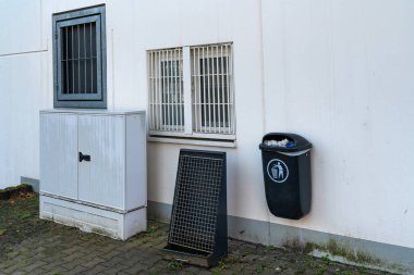 Beyaz bir dış duvara tutturulmuş siyah bir çöp kutusunun yanında gri bir depo var. Metal ızgaralı pencereler kentsel bir ortamda havalandırma ve güvenlik sağlar.