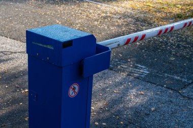 Otoparkın girişinde mavi bir giriş bariyeri var. Güvenlik kolu da var. Çevrede yaprakları dökülmüş bir kaldırım var..