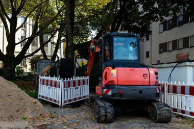 Bir şehir caddesinde kazı yapan bir kazıcı, kentsel alanda yakındaki ağaçların gölgesinin altında çitlerle çevrili bir inşaat alanının yakınlarını kazıyor..