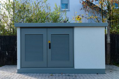 Şehir manzarasını geliştiren sessiz bir yerleşim yerindeki yemyeşil alanların ortasında yer alan asfalt bir alanda gri bir hizmet kutusu belirgin bir şekilde duruyor..