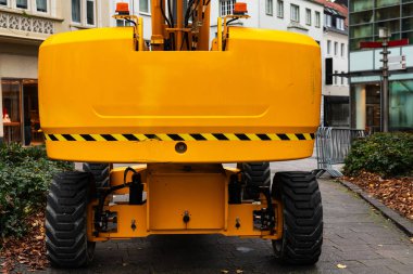 Şehir caddesinde büyük sarı bir inşaat makinesi var. Aracın tekerlekleri ve ayırt edici arka görüntüsü kentsel çevreler arasında belirgin bir şekilde gösteriliyor.