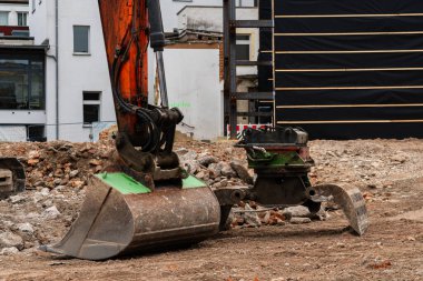 Bir kazıcı, bir inşaat sahasında özenle çalışarak şehir ortamında çakıl ve moloz taşır. Çevredeki binalar etkinliğe zemin hazırlıyor.