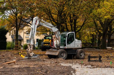 Yapı çalışmaları, yapraklarını ve ekipmanlarını etrafa saçan ağaçlarla çevrili renkli bir sonbahar manzarasında toprağı hareket ettiren bir kazıcıyla devam ediyor..