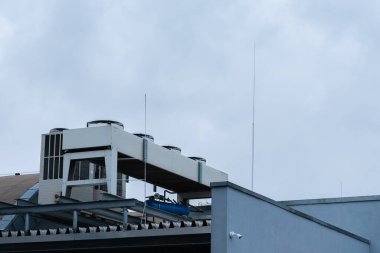 Çatıda, bulutlu bir gökyüzünün zeminine kurulmuş birden fazla ünitesi ve anteniyle kentsel sanayi manzarasını yakalayan bir soğutma sistemi bulunuyor..