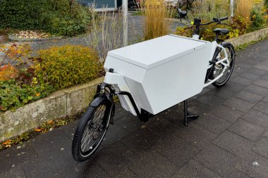 A cargo bike featuring a large white storage box stands parked along a city sidewalk surrounded by autumn foliage and greenery, showcasing urban transport solutions. clipart